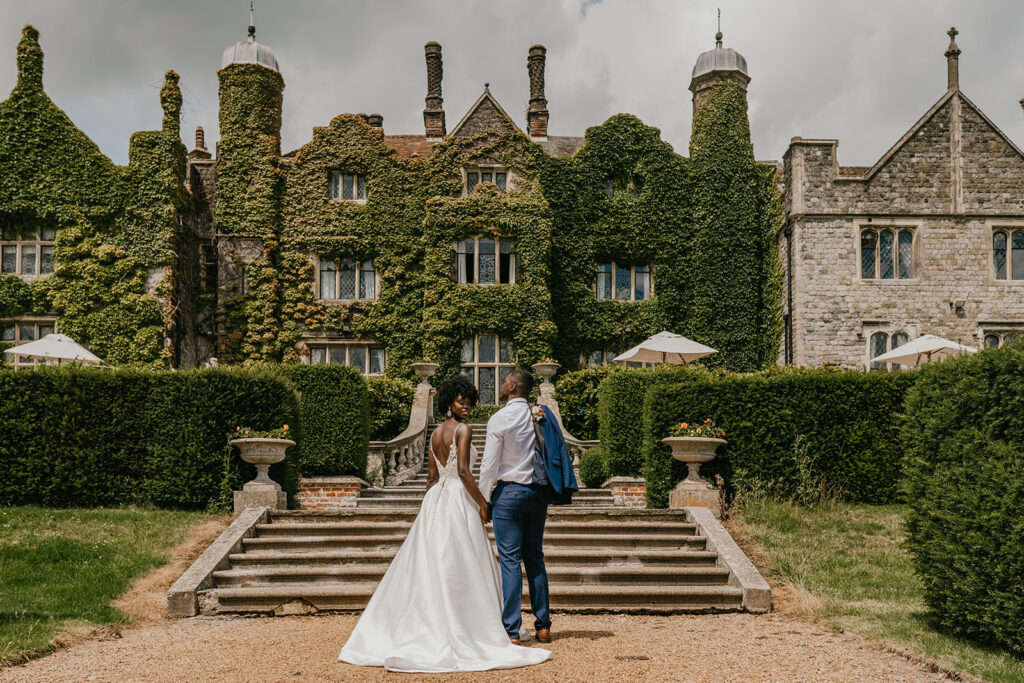 Newlywed couple walking toward an ivy-covered English manor house.