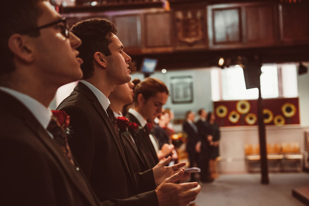 A couple exchanging vows in a bright church filled with stained-glass windows and floral arrangements.