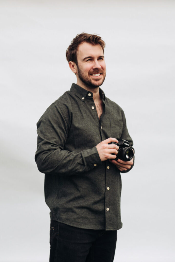 Portrait of wedding photographer Tom Keenan holding his camera, smiling naturally against a light backdrop