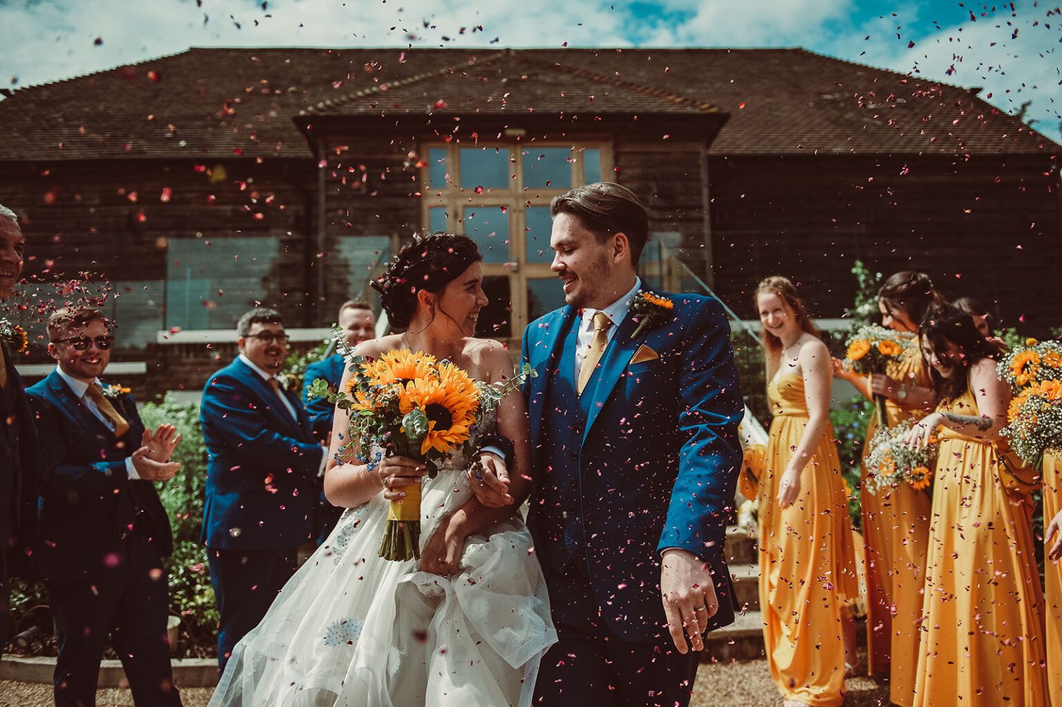 Bride and groom at Brookfield Barn having pink and red confetti thrown over them, the bride holds a bouquet of sunflowers, the bridesmaids in yellow and groomsmen in blue suits with yellow ties