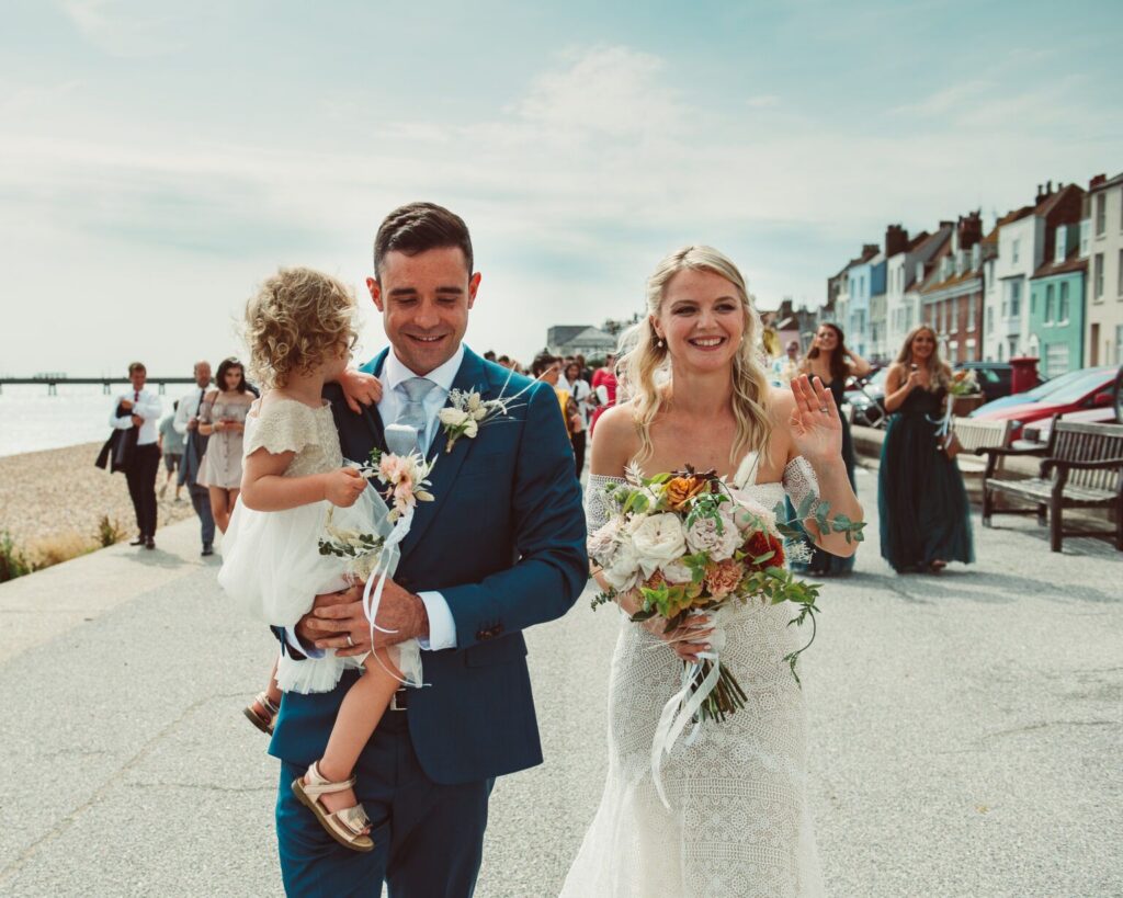 Bride and groom holding their daughter walking along the seafront in Deal as their bridal party and guests follow them along the promenade