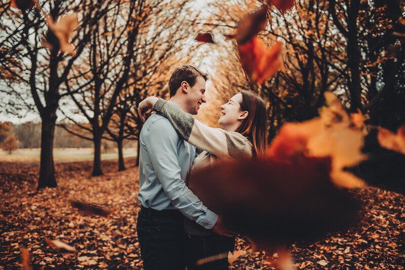 Couple embracing in a scenic autumn forest, surrounded by falling leaves during their engagement photoshoot.