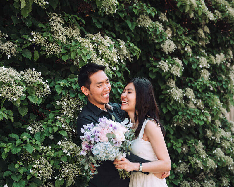 Happy engaged couple sharing a laugh in front of a lush, green floral backdrop