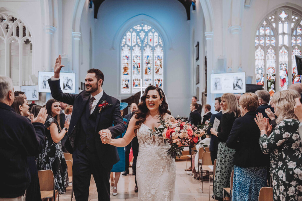 Luxury wedding at a church in Cambridge, bride is holding a bouquet of flowers, groom is being high-fived